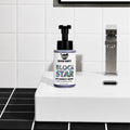 A bottle of 1 Hour Block Party Disruptor Anti-Humidity Mouse spray with a black pump and white label, placed on a white ceramic sink in a modern bathroom with black and white tiles.