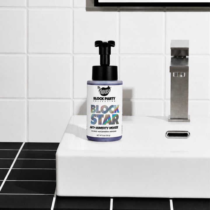 A bottle of 1 Hour Block Party Disruptor Anti-Humidity Mouse spray with a black pump and white label, placed on a white ceramic sink in a modern bathroom with black and white tiles.